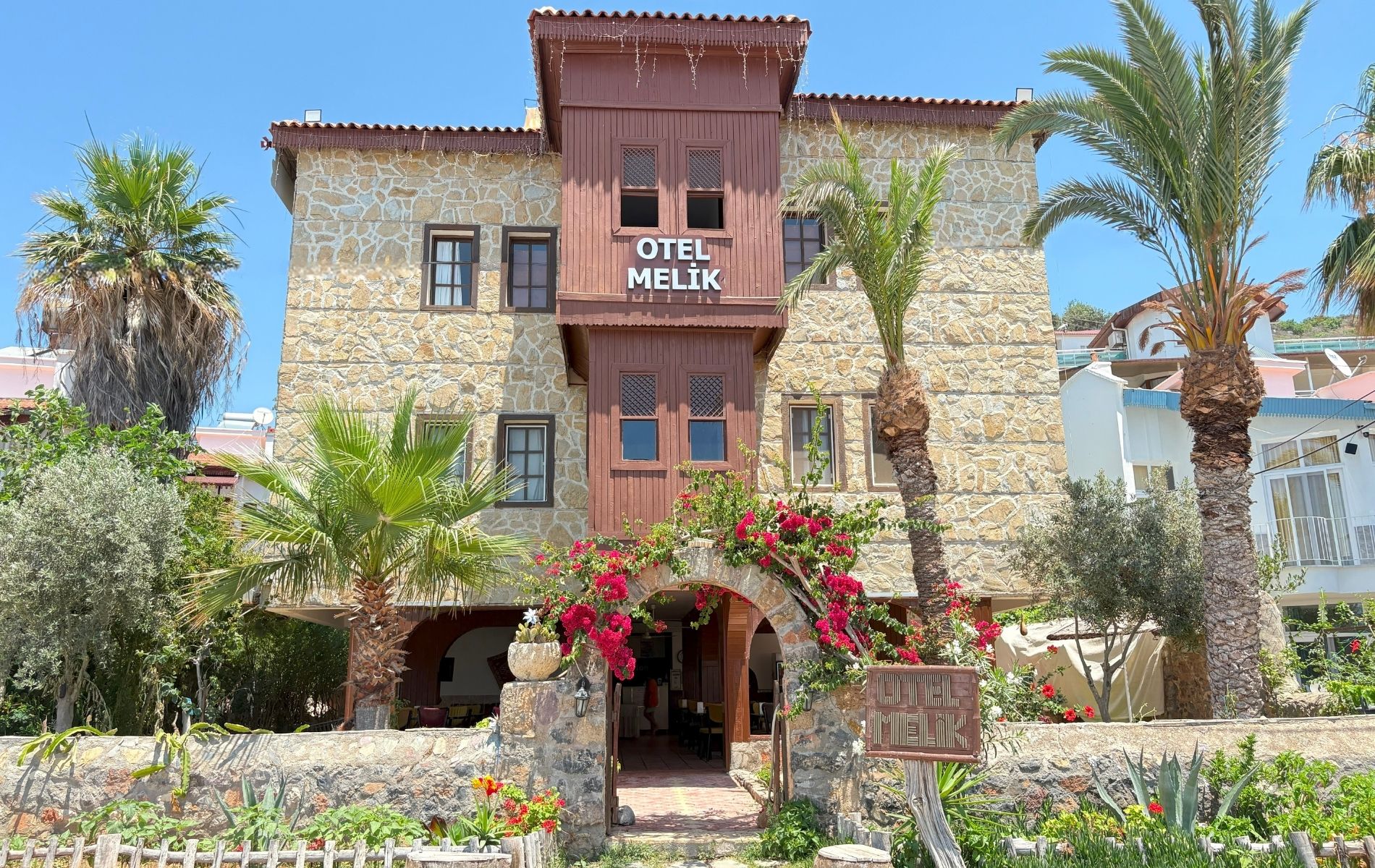 Otel Melik'in doğal taş ve ahşap cephesi, palmiyeleri ve çiçekli girişi